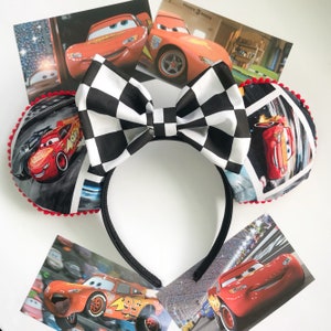May include: Black headband with a large checkered bow and two round ears featuring images of the animated character Lightning McQueen from the movie Cars. The ears are trimmed with red pom-poms. Several images of Lightning McQueen are scattered around the headband.