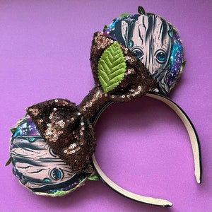 May include: Brown and green sequined Minnie Mouse ears with a fabric bow and a printed image of a character from the Guardians of the Galaxy movie. The character is a tree-like humanoid with a green leaf on its head.