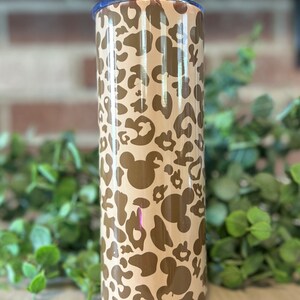 May include: A brown and tan stainless steel tumbler with a leopard print and Mickey Mouse silhouettes. The tumbler has a straw and a lid.