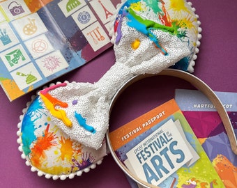 May include: White headband with a white bow and colorful paint splatter design. The headband has a white bow with sequins and a colorful paint splatter design on the bow and the headband. The headband is on top of a brochure that says "Epcot International Festival of the Arts" and "Festival Passport".