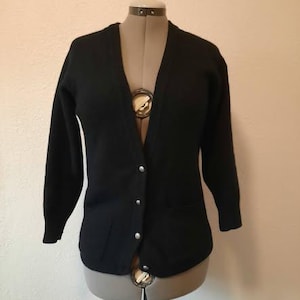 May include: A black cardigan sweater with a V-neck and long sleeves. The sweater has four silver buttons down the front and two pockets. The garment is displayed on a mannequin against a neutral background.