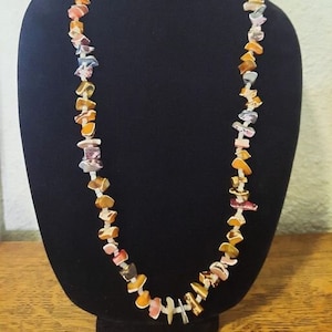 May include: A long necklace made of colorful, irregular shaped stones. The stones are mostly shades of orange, yellow, and pink, with some white and gray accents. The necklace is strung on a thin gold chain.