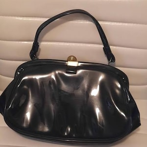May include: A glossy black handbag with a curved handle and a gold-colored clasp. The vintage purse has a rounded shape and a shiny, reflective surface. The handle is attached to the top of the bag with small metal attachments.