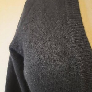 May include: Close-up of a black V-neck sweater. The sweater is made of a soft, textured material. The image focuses on the shoulder and upper chest area, highlighting the fabric's weave and the garment's construction.