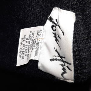 May include: Close-up of a black garment with a white tag. The tag reads "Jom Him" in a stylized script and lists the fabric composition as 70% lambswool, 20% other, and 10% nylon. The tag also indicates the garment was made in Hong Kong.