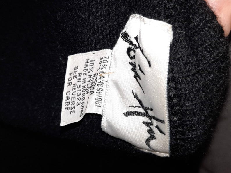 May include: Close-up of a black garment with a white tag. The tag reads "Jom Him" in a stylized script and lists the fabric composition as 70% lambswool, 20% other, and 10% nylon. The tag also indicates the garment was made in Hong Kong.