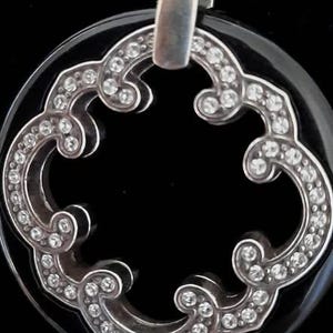 Pode incluir: Um pingente de &ocirc;nix preto com um design de filigrana prateada e detalhes de cristal transparente. O pingente tem a forma de um trevo de quatro folhas.