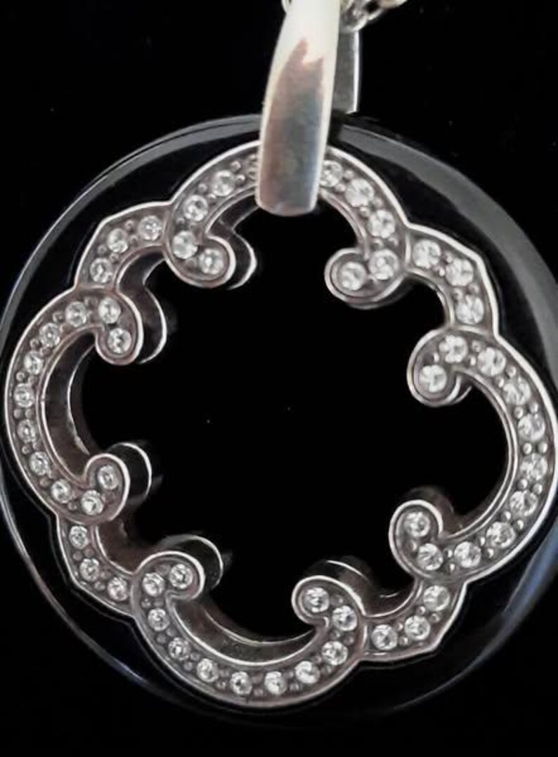 Pode incluir: Um pingente de &ocirc;nix preto com um design de filigrana prateada e detalhes de cristal transparente. O pingente tem a forma de um trevo de quatro folhas.