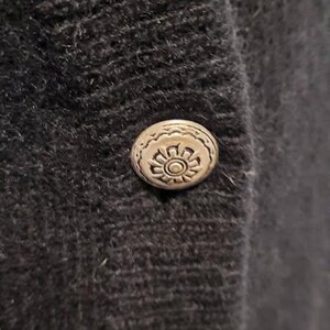 May include: Close-up of a dark gray, textured fabric with a decorative silver button. The button has a detailed floral design in the center and a scalloped edge. The button is attached to the fabric.