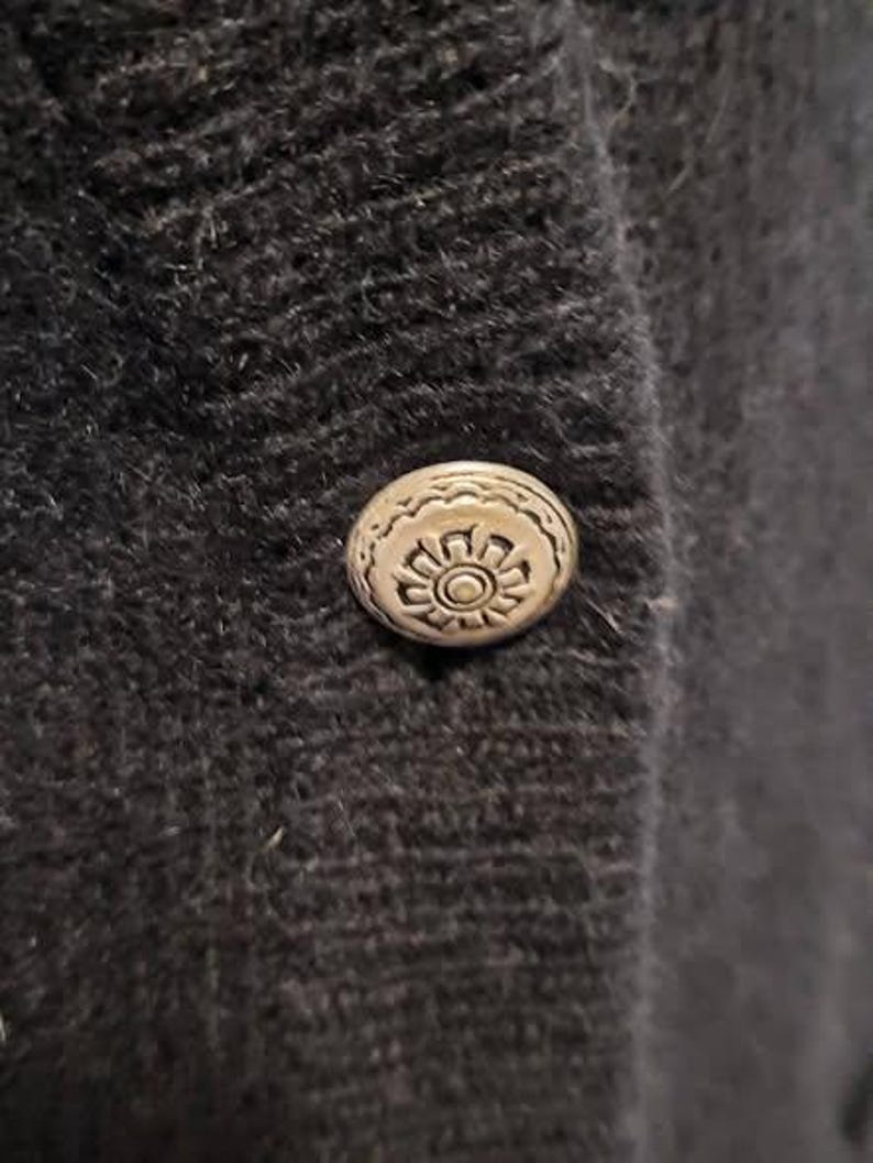 May include: Close-up of a dark gray, textured fabric with a decorative silver button. The button has a detailed floral design in the center and a scalloped edge. The button is attached to the fabric.