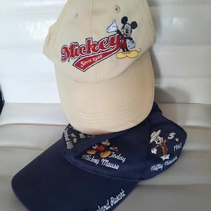 May include: Two baseball caps. The top cap is beige with a Mickey Mouse graphic and the word "Mickey" in red. The bottom cap is navy blue with "Disneyland Resort" and a Mickey Mouse graphic.