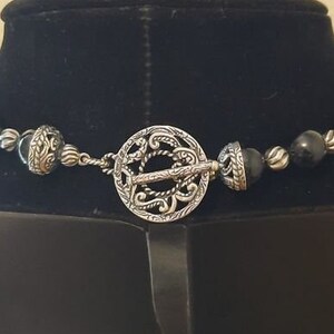 Peut inclure: Un collier de perles noires avec un fermoir argent&eacute; orn&eacute; d'un motif complexe. Le fermoir est un cercle avec un motif d&eacute;taill&eacute;.