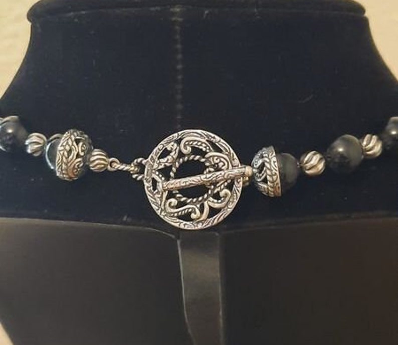 Peut inclure: Un collier de perles noires avec un fermoir argent&eacute; orn&eacute; d'un motif complexe. Le fermoir est un cercle avec un motif d&eacute;taill&eacute;.