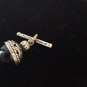 Peut inclure: Gros plan sur un fermoir argent&eacute; avec une perle d'onyx noire sur un collier. Le fermoir est orn&eacute; d'un motif d&eacute;taill&eacute; et est s&eacute;curis&eacute; par une petite barre.