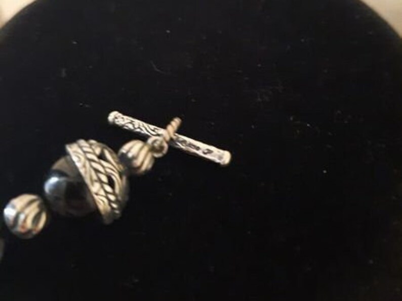 Peut inclure: Gros plan sur un fermoir argent&eacute; avec une perle d'onyx noire sur un collier. Le fermoir est orn&eacute; d'un motif d&eacute;taill&eacute; et est s&eacute;curis&eacute; par une petite barre.