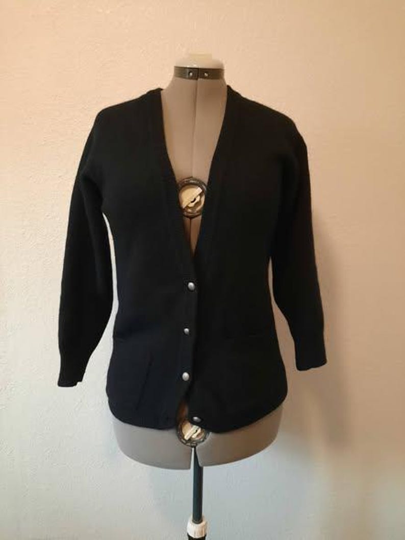 May include: A black cardigan sweater with a V-neck and long sleeves. The sweater has four silver buttons down the front and two pockets. The garment is displayed on a mannequin against a neutral background.