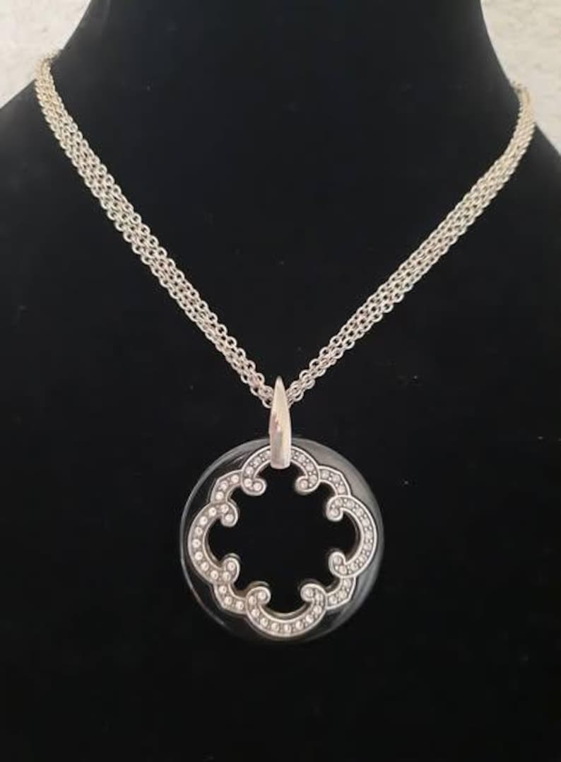 Pode incluir: Um colar de corrente de prata com um pingente preto e prateado. O pingente &eacute; de forma redonda e vazada com um desenho de trevo de quatro folhas.