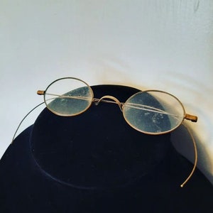 May include: A pair of round, gold-rimmed eyeglasses with clear lenses. The glasses are on a black surface.