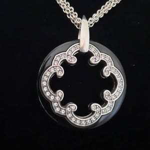 Pode incluir: Um colar de corrente de prata com um pingente preto que apresenta um design prateado em forma de diamante com um centro recortado.