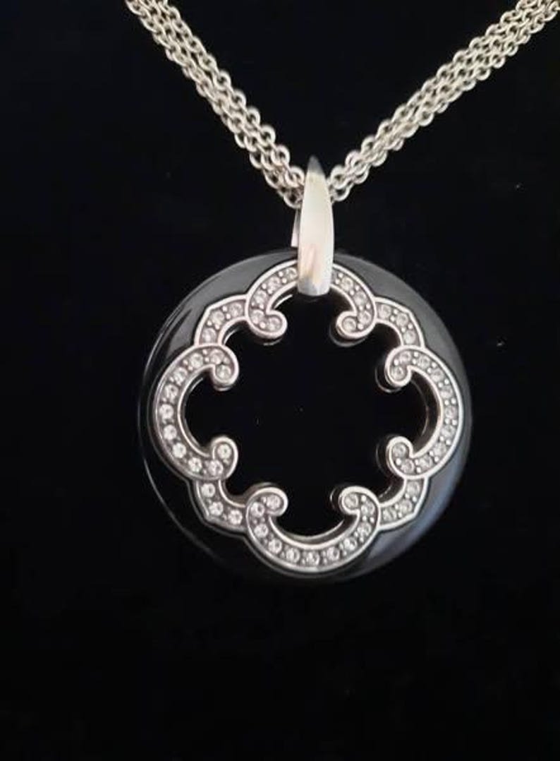 Pode incluir: Um colar de corrente de prata com um pingente preto que apresenta um design prateado em forma de diamante com um centro recortado.