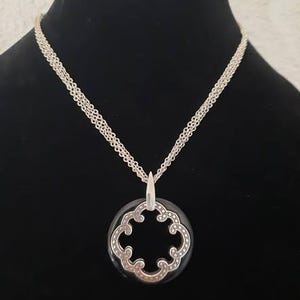 Pode incluir: Um colar de corrente de prata com um pingente preto e prateado com um design decorativo vazado. O pingente tem a forma de um trevo de quatro folhas.