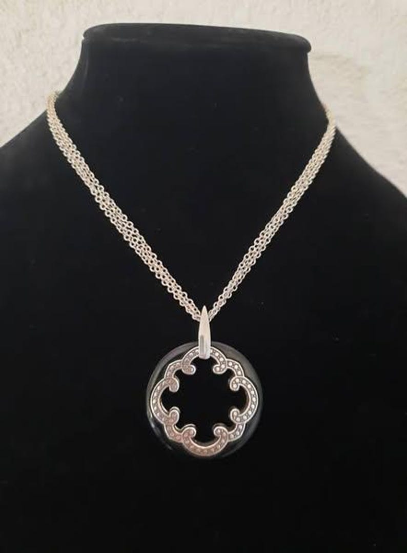 Pode incluir: Um colar de corrente de prata com um pingente preto e prateado com um design decorativo vazado. O pingente tem a forma de um trevo de quatro folhas.