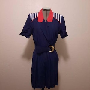 Puede incluir: Un vestido azul marino con cuello rojo y detalles de rayas blancas en los hombros. El vestido tiene mangas cortas y un cinturón a juego con una hebilla dorada. El vestido se exhibe en un maniquí.