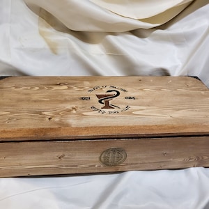 May include: A wooden box with a hinged lid. The lid has a circular design with a snake wrapped around a cup, and the text "001 024". The box has a brass latch and hinges.