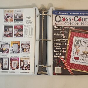 Könnte beinhalten: Ein blauer Ordner, der geöffnet ist, um Kreuzstichmagazine und -muster zu zeigen. Die rechte Seite zeigt ein Magazin mit dem Titel "Cross-Country Stitching" mit Weihnachtsthema. Die linke Seite zeigt verschiedene Kreuzstichmusterhefte.