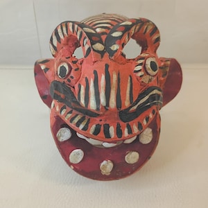 May include: A carved wooden mask with a red base color, featuring black, white, and cream accents. The mask has large eye openings, a wide mouth with white teeth, and a detailed, textured surface. The mask's design suggests a cultural or ceremonial purpose.