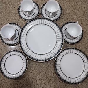 May include: A set of white porcelain dinnerware with a black and gold trim. The set includes a large dinner plate, a smaller salad plate, and a teacup and saucer.