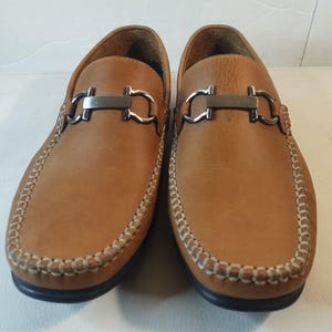 May include: A pair of brown leather loafers with a silver buckle detail. The loafers have a stitched edge and a black sole.