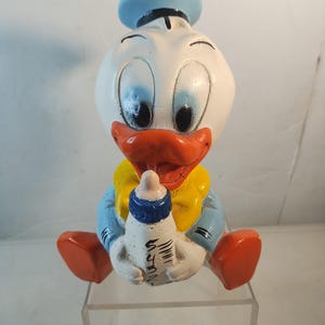 May include: A ceramic figurine of a baby duck wearing a blue sailor hat and holding a white baby bottle. The duck is sitting down with its legs outstretched and has a yellow bib. The figurine is painted in bright colors, including blue, white, yellow, orange, and black.