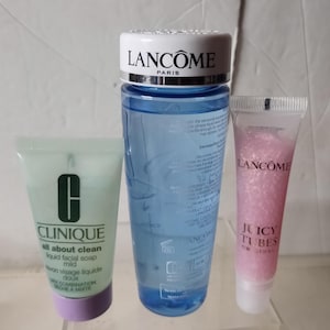 Puede incluir: Tres productos para el cuidado de la piel: una botella azul de Lancôme Tonique Douceur, un tubo verde de Clinique All About Clean Liquid Facial Soap Mild y un tubo rosa de Lancôme Juicy Tubes Original Lip Gloss.