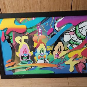 Op de afbeelding: Een ingelijst kunstwerk met stripfiguren met overdreven uitdrukkingen. Het levendige stuk gebruikt een palet van blauw, geel, roze en groen. Het zwarte frame omringt de kleurrijke illustratie en creëert een opvallend visueel contrast.