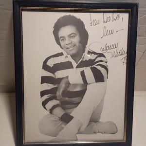 May include: Framed black and white photograph of a man wearing a striped shirt and white trousers. The photograph is signed "Johnny Mathis" and dated '78. The frame is black with a gold inner edge.