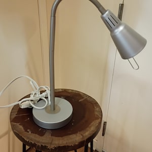 Puede incluir: Una lámpara de escritorio plateada con un brazo flexible de cuello de cisne y una pantalla cónica. La lámpara está sobre una base redonda plateada y se apoya en un taburete de madera. El taburete tiene una parte superior marrón oscuro y patas de metal negro.