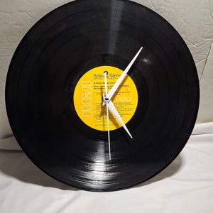 Puede incluir: Un reloj de pared hecho de un disco de vinilo negro con una etiqueta amarilla de RCA Victor en el centro. La etiqueta muestra texto que incluye Side 2, Stereo y A Warm Shade of Ivory de Henry Mancini and His Orchestra. Tres manecillas blancas están fijadas en el centro.