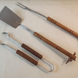 Puede incluir: Un juego de cuatro utensilios de barbacoa de acero inoxidable con mangos de madera. El juego incluye una espátula, pinzas y un tenedor de dos puntas. Los mangos de madera son de color marrón medio y tienen un lazo de cuero para colgar.