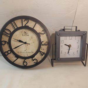 May include: Two clocks: a large, round wall clock with a dark brown frame and a smaller, square table clock. The wall clock has a cream-colored face with black numerals and hands. The table clock is gray with a white face and black numerals.