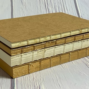 Puede incluir: Una pila de cuatro cuadernos con cubiertas de diferentes colores. El cuaderno superior tiene una cubierta marrón, el segundo cuaderno tiene una cubierta dorada, el tercer cuaderno tiene una cubierta blanca y el cuaderno inferior tiene una cubierta dorada. Los cuadernos están todos encuadernados con costuras decorativas.