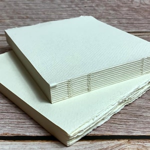 Puede incluir: Dos libros hechos a mano, uno con una cubierta blanca y el otro con una cubierta crema. El libro blanco tiene una encuadernación cosida visible. Ambos libros están hechos de papel.