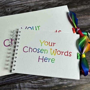 May include: Two white spiral-bound notebooks with the text "Your Chosen Words Here" in rainbow colors. A colorful ribbon is tied around the smaller notebook. The notebooks are on a wooden surface.