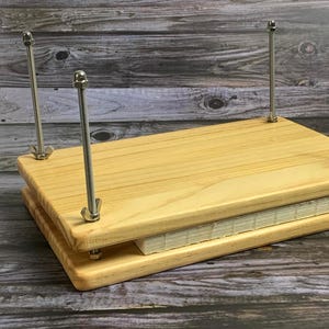 Puede incluir: Una prensa de libros de madera con cuatro tornillos de metal y una pila de papel en su interior. La prensa está hecha de madera clara y tiene un diseño sencillo.