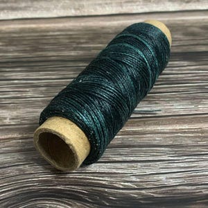 May include: A spool of dark teal waxed thread. The thread is tightly wound around a cardboard core. The color of the thread has a slight sheen, and the texture appears smooth. This thread is suitable for various crafting projects.