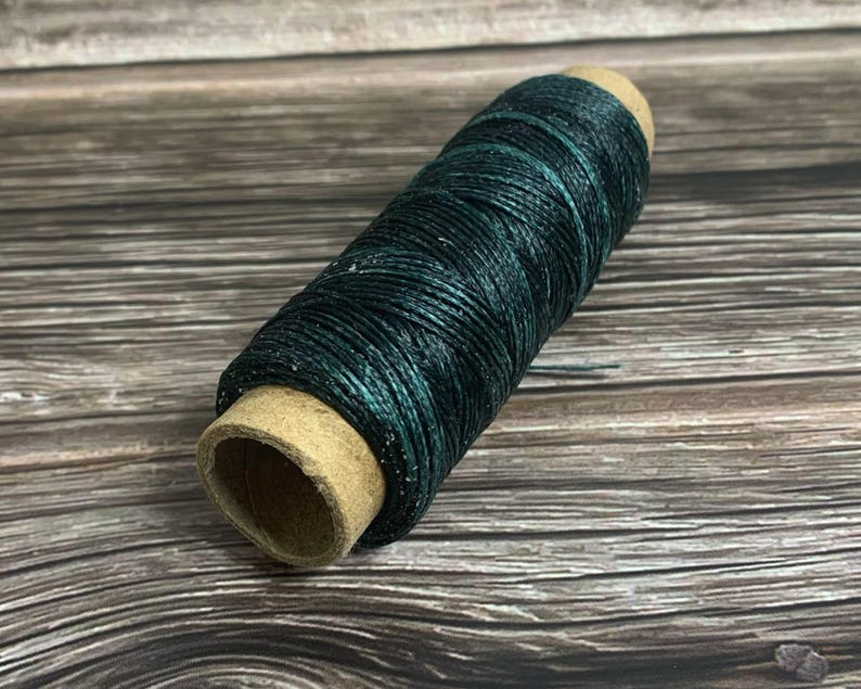 May include: A spool of dark teal waxed thread. The thread is tightly wound around a cardboard core. The color of the thread has a slight sheen, and the texture appears smooth. This thread is suitable for various crafting projects.