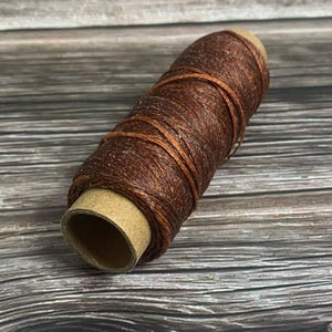 May include: A spool of brown waxed thread, ideal for leathercraft and other crafting projects. The thread is tightly wound around a cardboard core, showcasing its texture and color. The background is a wooden surface.