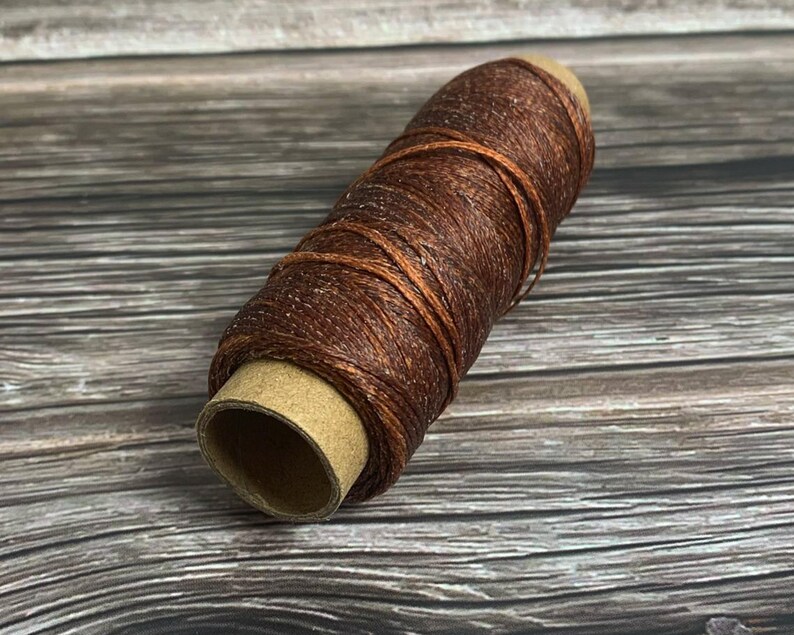 May include: A spool of brown waxed thread, ideal for leathercraft and other crafting projects. The thread is tightly wound around a cardboard core, showcasing its texture and color. The background is a wooden surface.