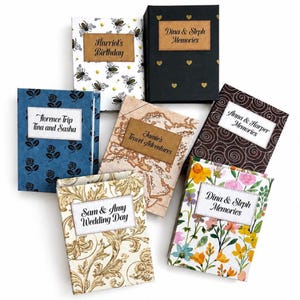 Peut inclure: Plusieurs petits livres décoratifs aux couvertures variées. Les motifs incluent des abeilles, des cœurs, des motifs floraux et des cartes. Chaque livre porte une étiquette avec du texte comme "Harriot's Birthday" et "Dina & Steph Memories".