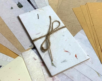 May include: A stack of handmade paper sheets with a natural, speckled texture. The top sheet is tied with a brown twine bow. The paper is a light cream color with flecks of brown, white, and red.